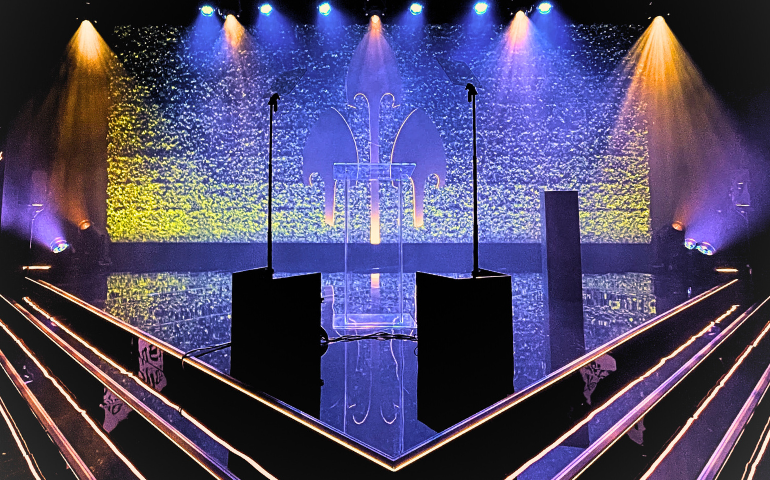 A shiny black pointed stage with 2 microphones. Blue and orange lights light up the back of the stage and the British Arrows logo.