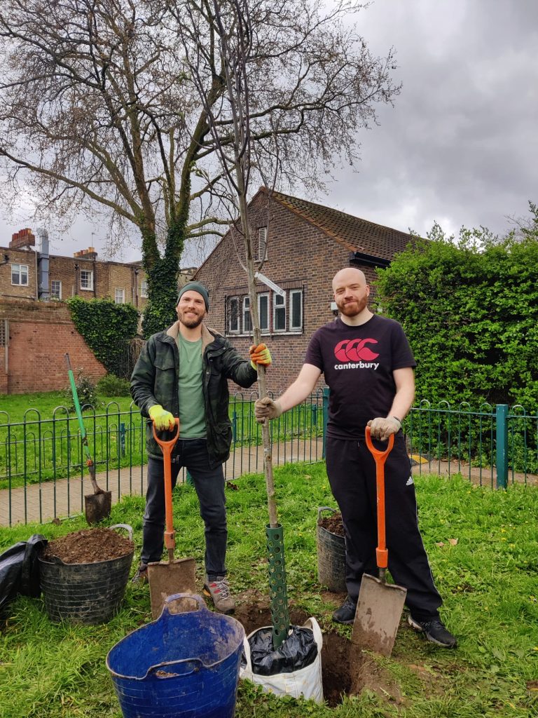 Stuart and Dave (copy staff) outdoors holding a tree upright in front of a hole they dug for it, with garden spades in their other hands, smiling 