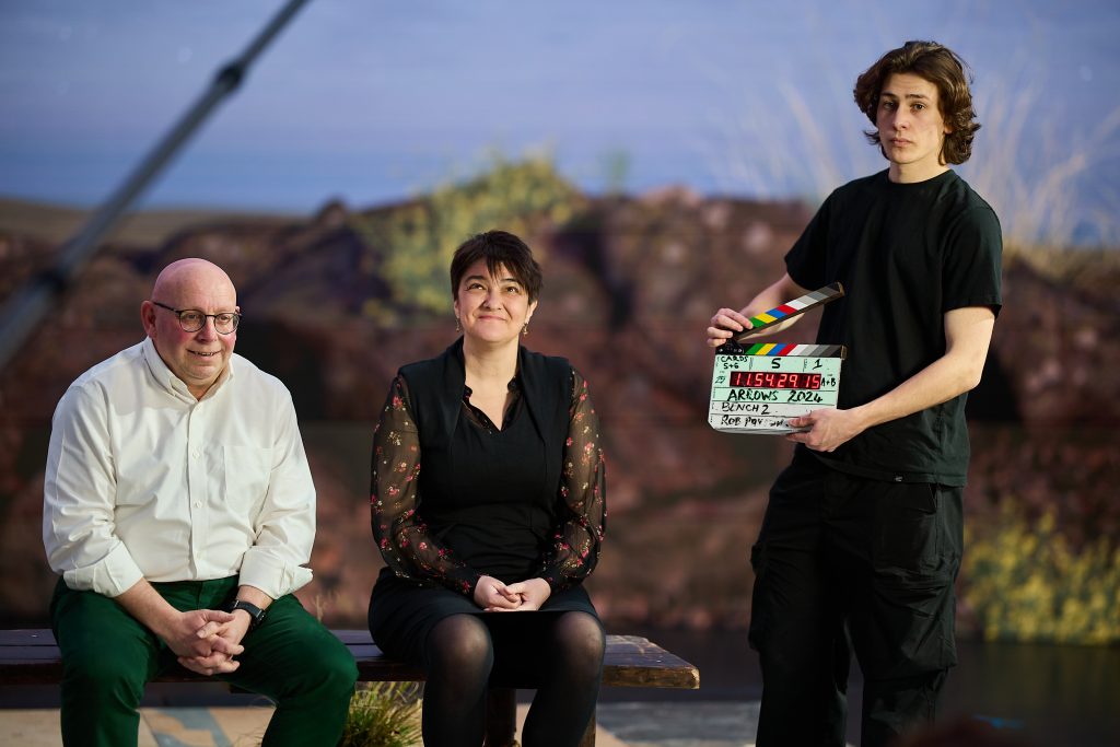 A man in a white shirt and a woman in a black dress sit on a bench in front of some greenery. A man in a black t-shirt stands next to them with a clapperboard.