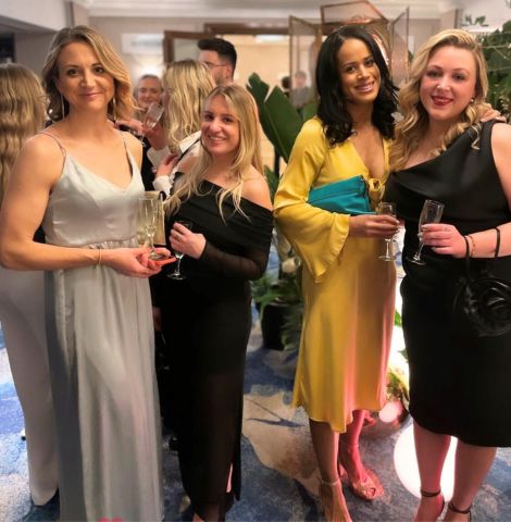 4 women in evening dresses stand and smile at the camera while holding champagne flutes