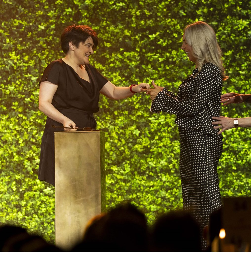 A smiling woman in a black dress stands by a podium. She hands an arrow award to another smiling woman who wears a black and white spotty dress.